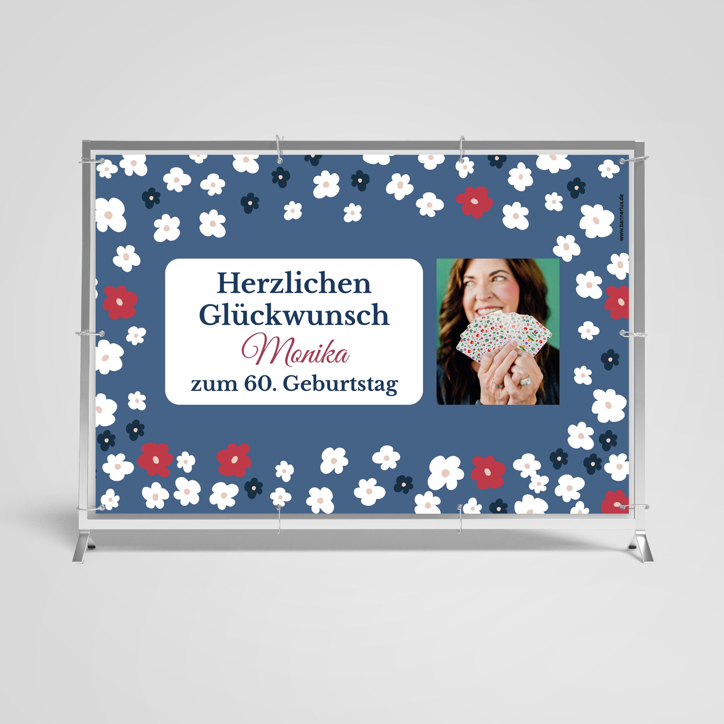 Feierliches Geburtstagsbanner zum 60. Geburtstag von Monika, gestaltet in einem dunkelblauen Hintergrund mit einem Muster aus roten, weißen und blauen Blumen. Ein Porträt von Monika, die lachend ein Stück Kuchen hält, ist in einem weißen Rahmen platziert. Der Text 'Herzlichen Glückwunsch Monika zum 60. Geburtstag' ist elegant in Weiß und Rot darüber und darunter angeordnet.