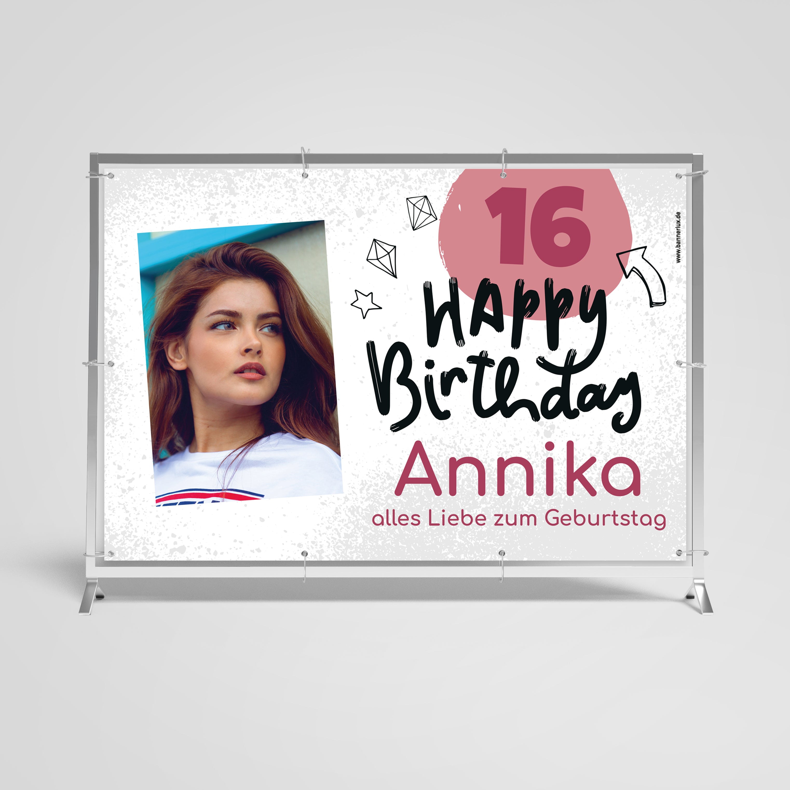 Geburtstagsbanner für den 16. Geburtstag von Annika. Weißer Hintergrund mit rosa Akzenten. Links ein Foto einer jungen Frau mit langen roten Haaren in einem weißen Oberteil. Rechts steht in großer Schrift 'Happy Birthday' und darunter 'Annika'. Oben rechts ist die Zahl '16' in einem rosa Kreis. Dekorative Elemente wie Sterne und Diamanten umgeben den Text. Unten steht 'alles Liebe zum Geburtstag'. Der Banner ist in einem Metallrahmen montiert.