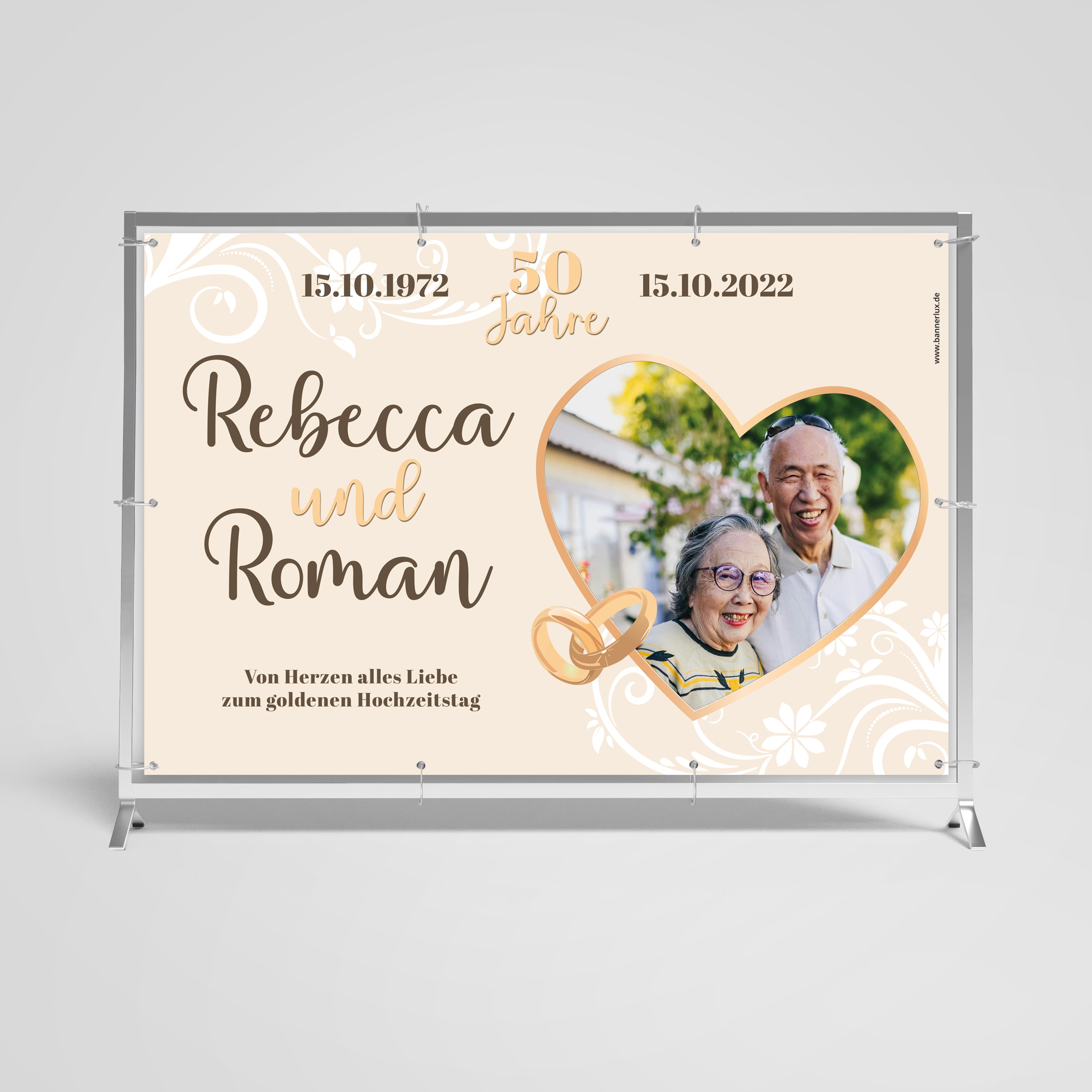 Personalisiertes Banner zum goldenen Hochzeitstag für Rebecca und Roman, gefeiert am 15.10.2022. Das Banner zeigt ein fröhliches älteres Paar umrahmt von einem herzförmigen Bildrahmen, dekoriert mit goldenen Eheringen und floralen Verzierungen, unterstrichen von den Daten '15.10.1972 - 50 Jahre - 15.10.2022'. Der Hintergrund ist in sanften Cremetönen gehalten.