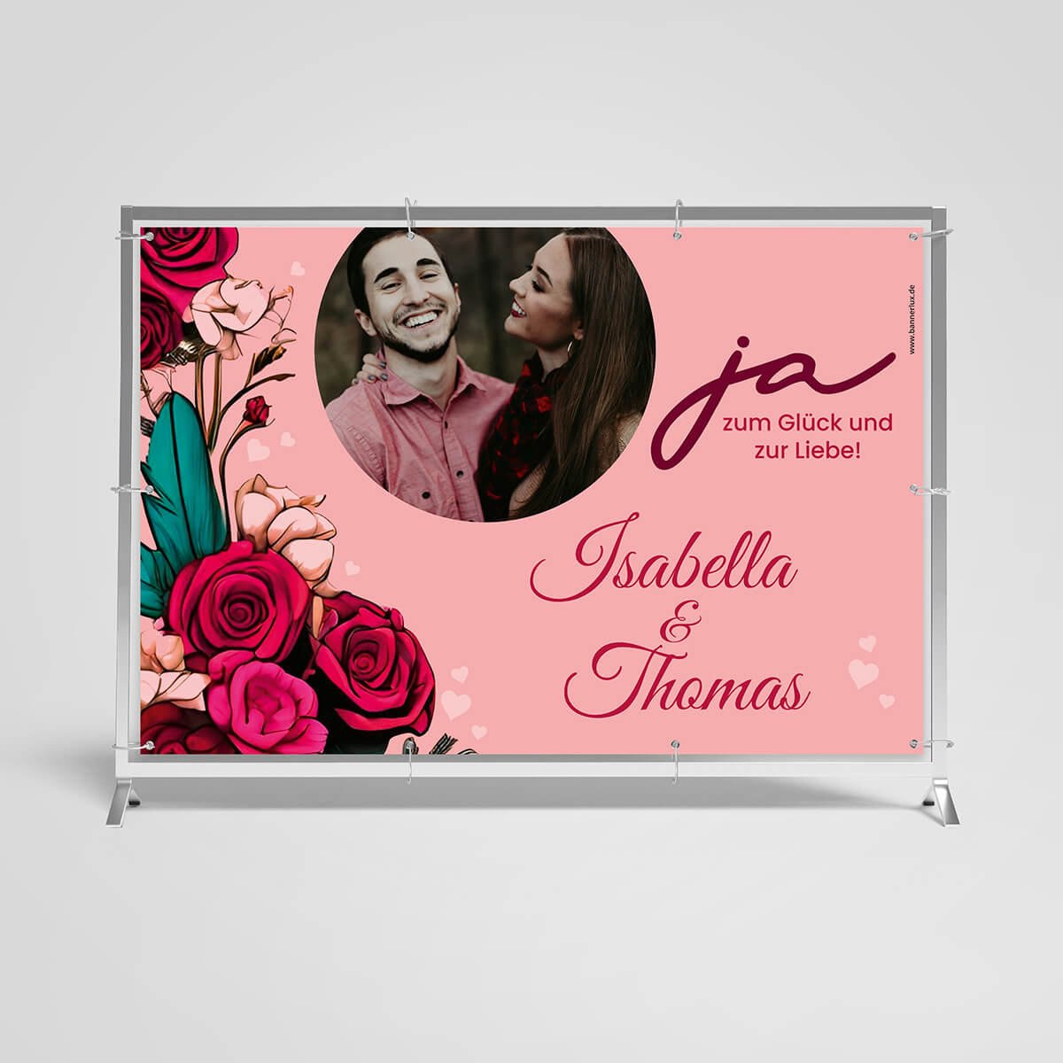 Personalisiertes Hochzeitsbanner in Rosa mit dem Namen 'Isabella & Thomas' und dem Schriftzug 'Ja zum Glück und zur Liebe!'. Das Banner zeigt ein lachendes Paar umgeben von einem Kreis aus lebhaften roten und rosa Rosen, isoliert auf weißem Hintergrund.